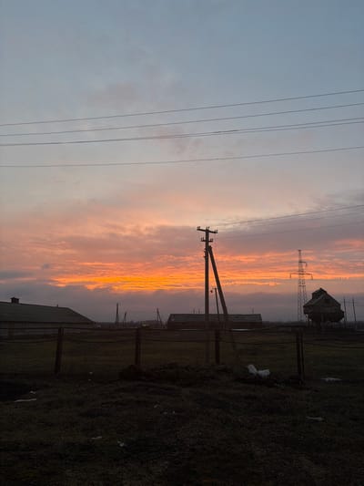The image displays a rural or semi-rural landscape in Bessergenovka, Russia, at either sunrise or sunset. The sky occupies the upper two-thirds of the frame, showing a gradient from light blue-grey at the top to intense orange and yellow along the horizon, indicative of low sun illumination. Stratocumulus clouds are present, notably brightly lit near the horizon. Multiple dark utility lines stretch horizontally across the sky.

The foreground consists of dark, sparse ground cover, primarily grass or weeds, appearing unkempt. A dark metal fence, possibly wire or chain-link, runs horizontally across the mid-ground. Behind the fence, a large clump of dark earth or debris is visible. To the right of center, a piece of white debris lies on the ground behind the fence.

In the mid-ground, several utility poles are prominent. A large wooden utility pole with multiple crossarms and insulators stands slightly left of center, angled towards the right. Additional smaller utility poles are visible in the distance. Silhouetted against the bright sky are several buildings: to the left, a long, dark, single-story structure with a dark green, gently sloped roof is visible. Further right, another dark, single-story building is present. On the far right, a tall, dark metal lattice transmission tower stands against the horizon. Next to this tower, a distinct dark structure, possibly an elevated grain silo or water tower with a complex, weathered shape, is silhouetted. No individuals, actions, or specific interactions are discernible within the scene.