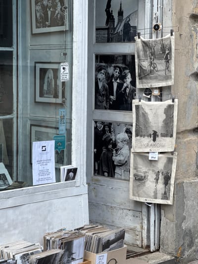The image shows the exterior of a shop in Kraków, Poland, located at the coordinates 50.053, 19.944. The shop is situated on a street corner and has a white facade. The window display features various photographs and postcards, including black and white portraits of individuals and scenes from the city of Kraków. Two signs are visible on the outside of the shop: one reads "NOTICE ALL ACTIVITIES MONITORED BY VIDEO SURVEILLANCE," and the other advertises "KRAKOW SOUVENIRS MADE WITH LOVE BY ...". Below the window display, there is a stack of postcards and photo albums organized for sale. Three cloth prints are hanging on a metal rack attached to the exterior wall, featuring black and white images of the city and its people. The image shows a street scene with people walking and a car driving by. The top print depicts a scene of people cycling through the streets, and the bottom print shows a man walking down a street. The prints all have the word "KRAKOW" displayed in a prominent location.