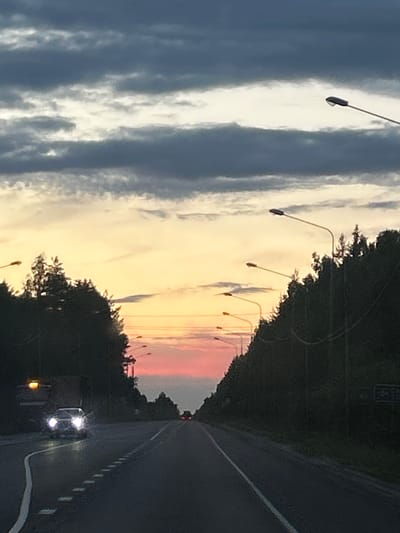 The image depicts a road scene at dusk, likely in Bel'skoe, Russia, with a dramatic sky transitioning from sunset colors to dark clouds. In the foreground, a dark asphalt road stretches into the distance, bordered by trees and a line of streetlights on the right. A white line marks the edge of the road on the left. A car with its headlights on is visible in the left lane, approaching the viewer. Another vehicle is further down the road. The sky above is filled with layered clouds, with a bright, yellowish-orange hue near the horizon, fading into pink and then dark grey clouds at the top. The overall impression is one of twilight on a rural or suburban highway.