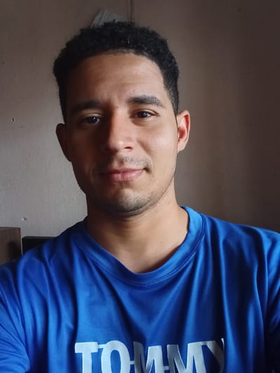 The image displays an adult male captured in a headshot within an indoor environment located in Duaca, Venezuela. The subject has a medium complexion, dark, closely cropped curly hair, and dark eyes. He possesses light stubble on his upper lip and chin, and maintains a neutral to slightly relaxed facial expression while looking directly forward. He is attired in a bright blue, short-sleeved t-shirt made of synthetic material, with white lettering partially visible on the lower chest, spelling out "TOMMY". The background consists of plain walls, appearing a light beige-brown on the left and a darker brown on the right. Illumination appears to be from the left, creating subtle shadows on the right side of the subject's face. No other individuals or prominent objects are distinctly visible in the immediate background.