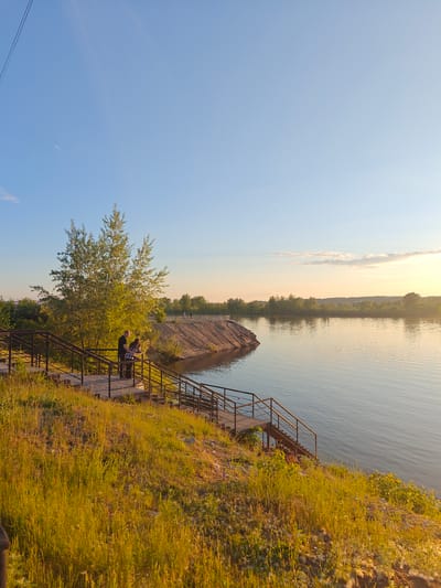 This image, captured in Nikolo-Beryozovka, Russia, depicts a serene riverside landscape under a clear blue sky transitioning to a warm, golden hue near the horizon, indicative of late afternoon or sunset. In the foreground, a grassy embankment covered in tall, dry-looking vegetation slopes downward. A multi-tiered wooden staircase with dark metal railings descends from the top of the embankment towards the river. Two individuals, one male in a dark shirt and one female in a plaid shirt, stand on an upper landing of these stairs, leaning against the railing and looking towards the water. Behind them, a large deciduous tree with bright green leaves is bathed in golden sunlight. The wide, calm river occupies the mid-ground, reflecting the sky and surrounding light, with gentle ripples visible on its surface. On the opposite bank, a treeline extends along the shore, with a distinctive sandy or earthen landform protruding into the water on the left. Several indistinct figures are visible on a raised path or structure along the far bank. White, fluffy detritus is scattered on the ground near the bottom right of the foreground.