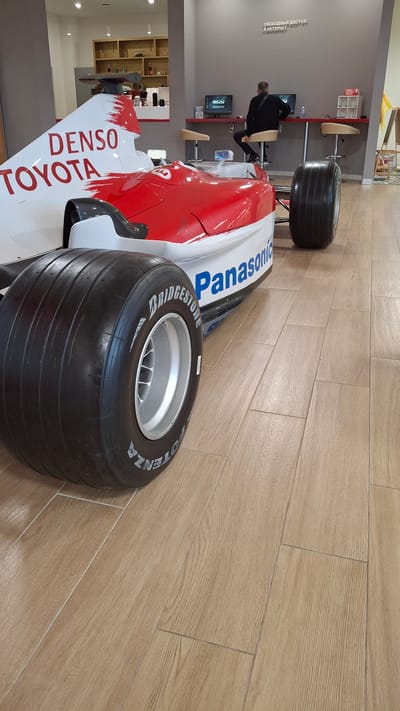 In Novogorelovo, Russia, a section of a red and white Formula 1 racing car is prominently displayed on a light brown tiled floor. The car features prominent "DENSO" and "TOYOTA" branding in red lettering on its white bodywork, and "Panasonic" in blue on its red side. A large, black racing tire with "BRIDGESTONE POTENZA" branding is visible in the foreground, mounted on a silver rim. In the background, a person wearing a black jacket is seated at a red counter, using one of two computer stations. The stations are equipped with monitors displaying the "ictv.ru" logo. The wall behind the counter has text that reads "СВОБОДНЫЙ ДОСТУП В ИНТЕРНЕТ ДОСТУП В ИНТЕРНЕТ" which translates to "FREE ACCESS TO THE INTERNET ACCESS TO THE INTERNET". The environment appears to be an indoor public space, possibly a showroom or a public internet access point.