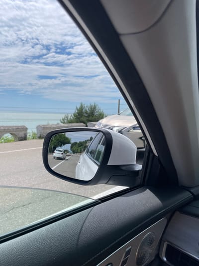 This is a close-up, landscape-oriented shot from inside a car, looking out through the side window and the side-view mirror. The camera is positioned to capture the interior of the car, including a portion of the dashboard with textured black leather and a speaker grill. The side-view mirror is a prominent subject, reflecting the scene behind the car.
The reflection in the mirror shows a white vehicle, likely a van or SUV, driving on a paved road. Trees and a cloudy blue sky are also visible in the reflection, suggesting an outdoor setting. The main part of the image, seen through the window, features the sky with scattered white clouds, and the edge of a body of water, possibly a sea or large lake. There are also some concrete structures and greenery visible along the roadside.
The image appears to have been taken during the daytime, with bright sunlight casting shadows and highlighting textures. The weather seems to be fair with clouds. The location context provided is Tsandrypsh, Abkhazia. There are no discernible people or text in the image. The overall impression is that of a car journey in a scenic, possibly coastal, location.