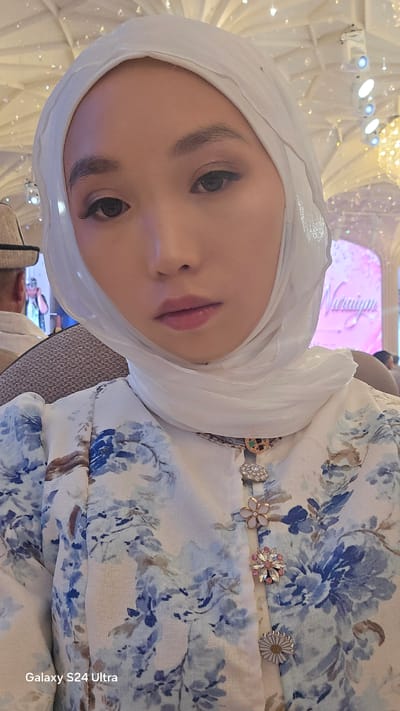 A young woman wearing a white hijab and a blue floral print dress is taking a selfie. She has subtle eye makeup on and her lips are painted a light pink. The background appears to be an indoor event, possibly a banquet hall, with elaborate lighting and decorations. A sign with pink lettering is visible in the background, though the text is largely illegible. The woman is seated, and a portion of another person's head and shoulder wearing a hat is visible on the left side of the frame. On the right side of the frame, blurry figures are present in the background. The image has a "Galaxy S24 Ultra" watermark in the lower left corner, suggesting it was taken with that device. The location is not specified in the image itself.