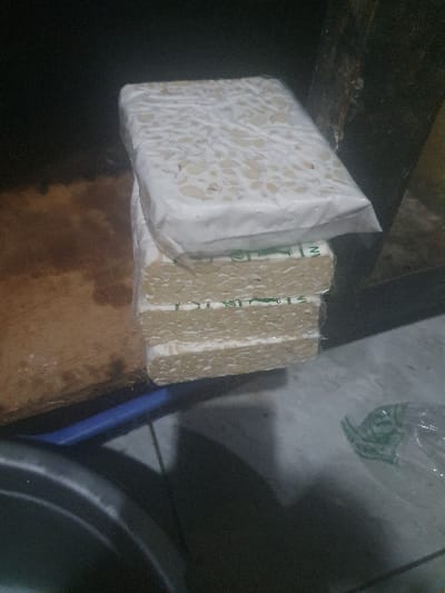 Three pieces of tempeh are stacked on top of each other on a countertop in Banguntapan, Indonesia. The tempeh is wrapped in clear plastic with green writing. The tempeh is resting on top of a brown surface. There is a blue surface below the countertop and a partially visible silver bowl to the left of the countertop. The scene appears to be indoors.