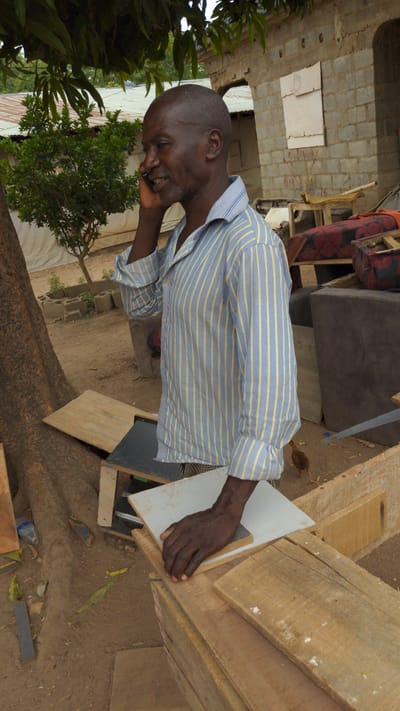 An adult man with dark skin, short hair, and a light beard is captured in an outdoor setting in Masaka, Nigeria. He is wearing a light blue and yellow striped long-sleeved shirt and is actively engaged in a phone call, holding a dark mobile phone to his right ear. He has a broad smile, indicating a pleasant conversation. His left hand rests on a light-colored wooden surface next to stacked wooden planks, suggesting he might be in a woodworking area or near unfinished furniture.
The immediate surroundings feature various wooden materials, including planks and what appear to be parts of unfinished structures or furniture pieces. A hand saw is visible near a dark cushion or sofa in the mid-ground. The ground is unpaved, consisting of dirt and scattered leaves, with a plastic bottle also visible. A small brown chick can be seen wandering on the ground near the wooden items.
In the background, there is an unfinished wall constructed from grey concrete blocks. Red graffiti-like text, "SAL-MAN," is written on one of these blocks. Lush green foliage from trees and bushes frames the left and central-right parts of the background. The scene appears to take place during the daytime, under natural light.