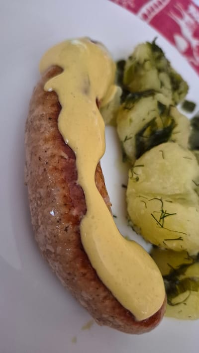 This is a close-up, overhead shot of a plate of food, likely a meal served in Krauja, Latvia. The main subject is a single, plump sausage, browned from cooking and generously topped with a creamy, yellow sauce, possibly mustard or hollandaise. The sausage rests on a white plate, partially obscuring a side dish of what appears to be potato salad. The potato salad is composed of roughly cut potatoes mixed with chopped green herbs, likely dill, and possibly a light dressing. A glimpse of a red and white patterned tablecloth is visible in the upper left corner, suggesting a home or casual dining setting. The lighting is bright and natural, indicating it was likely taken during the daytime. No people are visible, and the focus is solely on the food. The overall impression is one of a hearty and appetizing meal.
