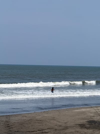 The image displays a single individual standing in the surf of a beach in Belalang, Indonesia. The person, viewed from the back, is shirtless and wears blue shorts, facing the open ocean. Breaking white waves surround the individual, whose lower legs are submerged in the water. The foreground features a dark, damp sand beach transitioning to drier sand. The midground shows the ocean with multiple wave lines, extending to a distant horizon under a clear, light blue sky.