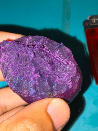 The image shows a close-up view of a person's hand holding a piece of a vibrant purple, crystalline mineral. The mineral has a rough, irregular surface with visible facets and some areas appearing to be a deeper, almost blueish purple. The background is a teal-colored surface, possibly a table or mat, with a shadow cast by an object at the top left, which appears to be a red lighter. The lighting suggests an indoor setting. The focus is primarily on the texture and color of the mineral, with the hand providing scale and context. The skin tone of the hand is medium-brown. There is no visible text in the image. The activity taking place is simply the holding and displaying of the mineral.