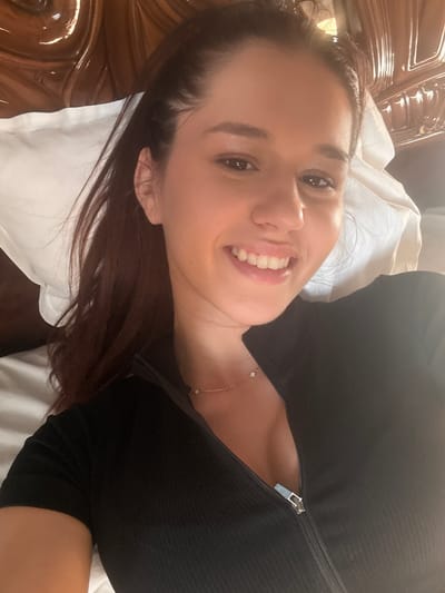 The image shows a woman lying on a bed, possibly indoors. She is smiling and looking towards the camera. She is wearing a black ribbed top with a zipper at the neck. A delicate necklace is visible on her neck. The background includes a white pillow and a wooden headboard. The location is not evident from the image; the assumption of Novo Zhelezare, Bulgaria, cannot be confirmed visually.