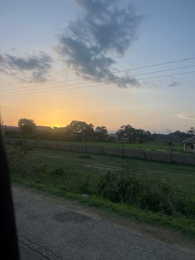 This photograph, taken in Rinjim Gani, Nigeria, depicts a sunset over a semi-rural landscape. The sun, a bright golden disc, is positioned low on the horizon on the left side of the frame, partially obscured by the silhouettes of structures and dense foliage. The sky exhibits a gradient, transitioning from a warm orange and yellow near the horizon on the left to a pale blue in the upper right quadrant, with scattered grey clouds spanning the upper half. Three distinct power lines run horizontally across the upper mid-ground. A small, dark, unidentified object, possibly a bird, is visible in the upper central-left sky.
In the mid-ground, a long, grey, cinder block wall extends horizontally across the scene, separating a wide expanse of green grassy field from the area beyond. Behind the wall and field, several large trees are prominent, and the roofs of some buildings are visible. On the far right, a solitary, small, yellow-painted building with a dark, sloped roof is situated near the edge of the grassy area. Distant hills or elevated terrain are present in the background on the right.
The immediate foreground features a dark, textured surface, indicative of a gravel or unpaved road, which runs diagonally from the lower left towards the lower right. Adjacent to the road on the right is a strip of dense green vegetation and bushes. A dark, vertical element is visible along the far left edge of the image, suggesting the photograph was taken from within a vehicle. No people are present in the scene.