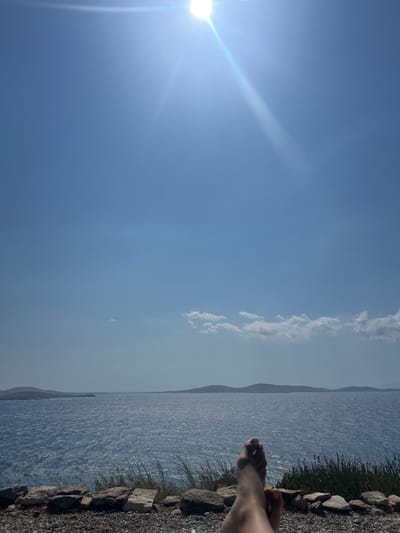 The scene is set in Agios Ioannis Diakoftis, Greece, overlooking a large body of water under a clear sky.
**Environment:** The upper two-thirds of the image display a bright, cloudless, light blue sky, with the sun positioned in the upper left quadrant, emitting a prominent lens flare and light rays. A horizontal band of small, white cumulus clouds is visible across the middle-right portion of the sky. Below the sky lies an expansive, deep blue sea with noticeable surface ripples and choppiness, reflecting sunlight. Along the distant horizon, multiple dark, undulating landmasses, consistent with islands or distant coastline, are discernible.
**Foreground:** The immediate foreground features a rocky and gravelly terrain, composed of various sized light grey and brown stones. Sparse, green, blade-like vegetation and low-lying bushes grow intermittently among the rocks, particularly on the right side of the frame.
**Subjects:** In the lower center-right, a pair of bare human feet are visible. They appear light-skinned and are positioned in a relaxed, overlapping manner, suggesting an individual is reclining or resting while observing the view.
**Actions/Interactions:** The singular action captured is the implied repose of the individual, indicated by the visible feet, within the natural coastal environment. No other individuals or interactions are present.