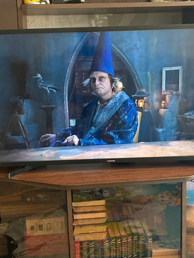 The image shows a television screen displaying a scene from a movie or show. A person wearing a blue pointed hat with white stars and a blue robe with a white patterned trim is seated at a table. Their hands are resting on the table. In the background, there is a blurry figure wearing a hat and holding a lantern, and a framed picture on the wall. To the left, a bird-like statue is visible. The television is placed on a wooden shelf, below which are stacked books and other items. The scene depicted on the screen has a fantastical or historical theme. The image does not contain any information or context about the city of Gornye Kljuchi, Russia.