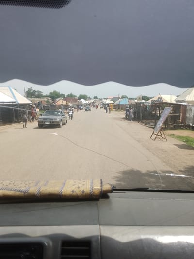 The image captures a street scene in Beli, Nigeria, likely during the daytime, given the illumination. The sky is dominated by a large, dark, and ominous cloud formation that spans the upper portion of the frame, with a distinctly wavy lower edge. This suggests impending or ongoing stormy weather.
The street is paved and appears to be a main thoroughfare in a town or village. On either side of the road are numerous small shops and market stalls, many with corrugated metal roofs. Some stalls have awnings or overhangs. The architecture suggests a local, possibly informal, commercial area.
Several people are visible, walking along the road and near the stalls. A few individuals are dressed in traditional attire, including flowing robes. There are also motorcycles and a car on the road, indicating regular traffic. One car, a sedan, is prominent in the foreground on the left side of the road. The presence of a few scattered trees indicates some vegetation in the area.
In the foreground, obscuring the bottom of the image, is the dashboard of a vehicle, with a textured material draped over a part of it. This suggests the photo was taken from inside a car, looking out through the windshield.
On the right side of the road, a wooden easel displays a sign with some text and imagery, possibly an advertisement or information board. The overall atmosphere is that of a busy, though potentially unsettled, day in a Nigerian community.
Visible text includes "LIMITED" on a structure in the background and partially visible text on the sign on the easel. The license plate on the car is also visible but unreadable.