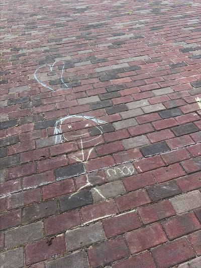 The image shows a brick-paved walkway with chalk drawings on it. In the upper left portion of the image, a blue chalk drawing of a heart is visible. Further down and to the right, there is a chalk drawing of a stick figure person with a round head, arms, and legs. The head has rudimentary facial features, including eyes and a smile. To the right of the stick figure, there are some chalk marks that appear to spell "moo 1". The walkway is paved with rectangular bricks of varying shades of red and gray. Some small green weeds are growing in the gaps between the bricks. The scene appears to be outdoors, likely in a public space. The location is identified as Rijswijk, Netherlands.