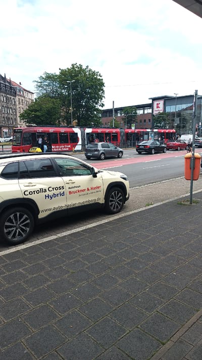 In Nuremberg, Germany, a beige Toyota Corolla Cross Hybrid, serving as a taxi for Autohaus Bruckner & Hasler, is parked on a brick sidewalk. The car displays advertising text, including its model, hybrid status, dealership name, and website address. In the background, a red tram with passengers travels on a street lined with a red traffic lane. A Kaufland supermarket, with its prominent red logo, is visible behind the tram. Other vehicles, including a grey Volkswagen Golf and a black sedan, are present on the road. The scene is set under a partly cloudy sky, with a trash can on a pole to the right of the frame.