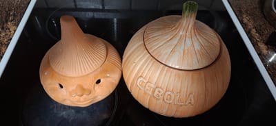 Two ceramic onion-shaped containers are placed on a black stovetop. The container on the left is smaller and has a face with two eyes and a mouth. The container on the right is larger and has the word "CEBOLA" inscribed on its side. The stovetop appears to be part of a kitchen. The surrounding countertop has a granite pattern.