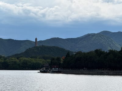 This forensic description details a wide landscape photograph taken in Haidian District, China, depicting a large body of water in the foreground with forested hills and architectural elements in the midground and background under an overcast sky.
The foreground is occupied by a calm, greyish-white body of water, showing fine ripples across its surface. In the midground, along the right shoreline, a long, ornamental stone balustrade or railing runs horizontally. Behind this balustrade, a dense line of dark green trees obscures much of the land, though indistinct figures of people are visible walking along a path or promenade. Further left along the shoreline, several small structures with red-tiled roofs and lighter-colored walls are situated at the water's edge, accompanied by a few small boats moored nearby.
The background consists of multiple layers of rolling hills, uniformly covered in dense green foliage. These hills recede into the distance, appearing progressively lighter due to atmospheric perspective. On the most prominent hill, positioned towards the left-center, a tall, multi-tiered pagoda with a distinct architectural style is visible. Another slender, antenna-like structure is faintly visible on a more distant hill to the right of the pagoda. The sky above is entirely overcast, dominated by a mix of light grey and blue-grey clouds.