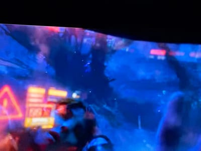 The image shows a blurry scene from what appears to be a video game or film. Dominant colors are blue and hints of red/orange. A blurred traffic sign with a triangular shape is visible on the left side, illuminated by orange light. Other blurred objects, possibly buildings or structures with red lights, are in the background. The foreground contains indistinct figures or objects, partially obscured by the overall blur. The scene is dark and details are unclear. It is not possible to determine any specific location within the city of El Bajío, Venezuela, due to the lack of clear identifying details.
