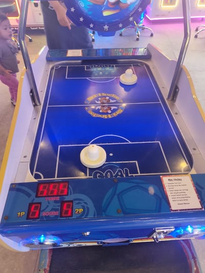 A low-angle medium shot captures an air hockey table in what appears to be an arcade or entertainment center. The table's surface is predominantly blue with white markings delineating the game area. Two white pucks are visible on the surface, one near the center and another at the bottom. The control panel at the front of the table features red digital displays showing "8.8.8." for TIME, "8" for 1P SCORE, and "8" for 2P SCORE. A small child, a girl with dark hair wearing a grey top and pink pants, is partially visible on the left side of the frame, looking towards the air hockey table. The background shows parts of other arcade machines with colorful lights, specifically pink and yellow hues. A sign with text, likely instructions for the game, is affixed to the right side of the table. The overall environment suggests a lively indoor entertainment setting. The location of Gunung Putri, Indonesia, is not ascertainable from the visual information provided in the image.
