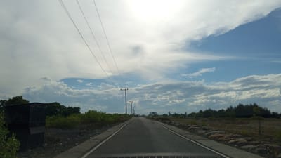 A straight asphalt road stretches into the distance under a partly cloudy sky in Pusong, Indonesia. The road is bordered on the left by a dark, weathered structure resembling a dumpster or storage bin, followed by lush green foliage and trees. On the right, the roadside consists of dry, sandy ground scattered with large rocks, and sparse vegetation. Multiple utility poles with overhead power lines are visible along the left side of the road, receding into the distance. A few vehicles can be seen on the road ahead, and a large, unfinished concrete structure is visible on the right horizon. The overall environment appears to be a rural or developing area.