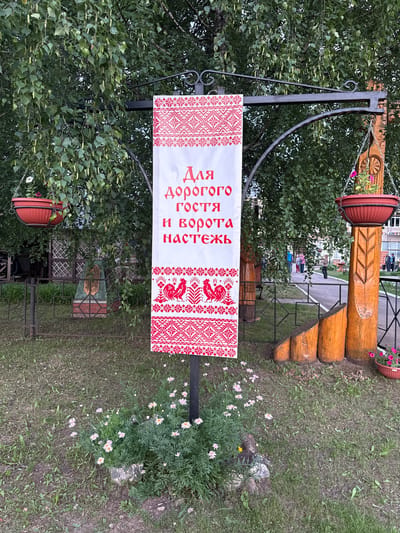 In the city of Kukui, Russia, a decorative banner hangs from a black metal archway, flanked by birch trees. The banner, primarily white with red embroidered details, features Cyrillic text that reads "Для дорогого гостя и ворота настежь" (For a dear guest, the gates are wide open). Below the text, two red roosters are embroidered, followed by intricate geometric patterns. Two red hanging planters, one with a white flower and another with pink flowers, are suspended from the archway. A wooden post with carved details stands to the right of the banner. The foreground is a grassy area with scattered daisies and some rocks. In the background, a fence encloses a yard with a checkered fence and a small monument. A pathway is visible, with several people walking in the distance.