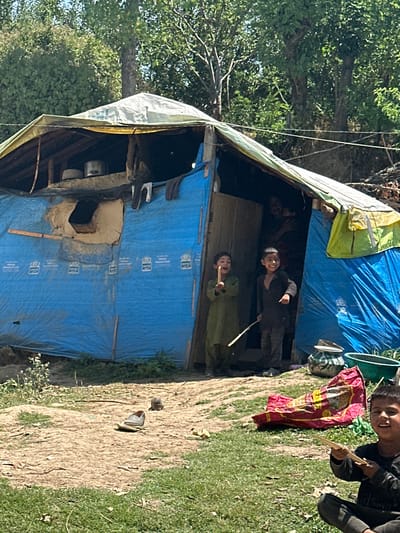 A group of children are playing in front of a makeshift shelter made of blue tarpaulin and a patched roof. The shelter appears to be in a rural or impoverished area, with dirt ground and overgrown grass. Inside the shelter, cooking pots are visible. One child in the foreground, looking directly at the camera, is holding a stick. Two other children are standing in the entrance of the shelter, one holding a stick as well. A bright red and yellow object, possibly a blanket or mat, lies on the ground near the shelter. The background consists of trees and foliage, suggesting an outdoor setting.