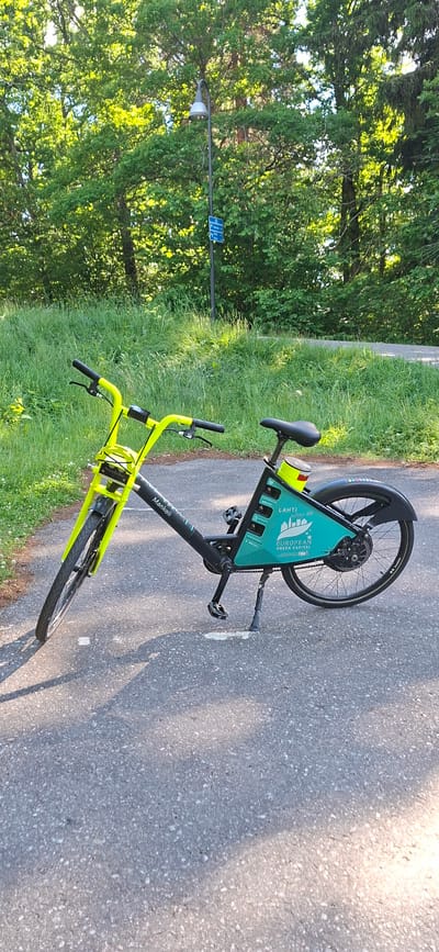 An electric bicycle with a lime green frame is parked on a paved asphalt path outdoors. The bicycle has "Mankeli" and "freebike" branding visible. The rear of the bicycle has a turquoise panel with "LAHTI" and "EUROPEAN GREEN CAPITAL 2021" text. The bicycle is in a park-like setting with lush green grass and mature trees in the background. A lamppost with a blue sign is visible behind the bicycle. The sun is casting shadows on the asphalt path.