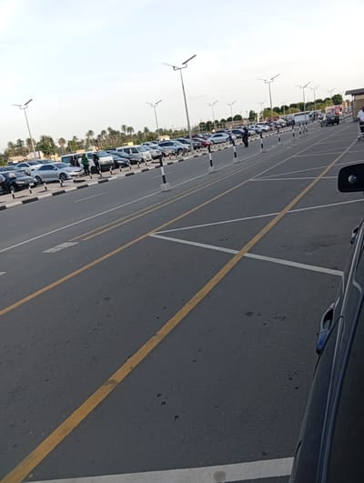 The image depicts an outdoor scene in what appears to be a parking lot adjacent to a road in Omagwa, Nigeria. The location is characterized by multiple lanes demarcated with yellow and white lines. Several cars are parked in an adjacent area lined with black and white bollards. In the background, there are palm trees. The presence of security personnel and parked cars suggests the scene is likely at an airport or a similar transport hub. A portion of a blue car is visible in the foreground on the right. The sky is overcast.