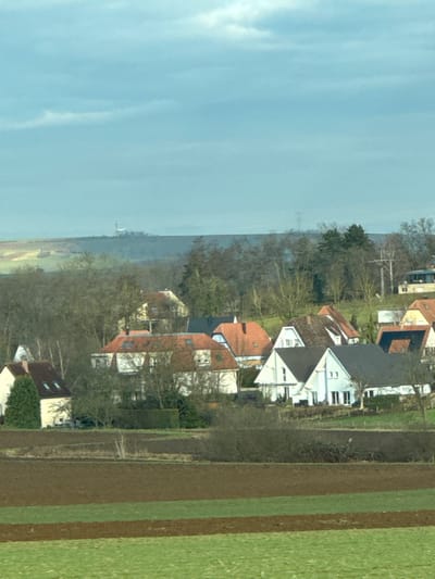 The image depicts a rural landscape in Hurtigheim, France, under an overcast sky. In the foreground, agricultural fields are visible, consisting of horizontal strips of green grass and freshly tilled brown earth. The midground features a cluster of residential houses, primarily two-story structures with varied roof designs in shades of terra cotta, dark grey, and light brown. Building facades are predominantly white, cream, or light beige. The houses are interspersed with numerous deciduous trees, mostly bare, suggesting winter or early spring, along with some evergreen trees and hedges. No individuals are visible. In the background, a distant rolling hill or ridge line is visible, with a prominent tall, slender white structure, possibly a church steeple or tower, situated on its peak. To the right of this structure, a faint power line pylon is discernible against the sky. The sky is light blue-grey with scattered clouds.