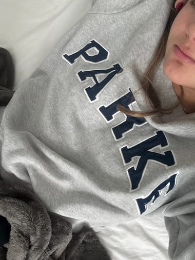 A person wearing a grey sweatshirt with the word "PARKE" in dark blue, with a white border around each letter, is lying on a white sheet. The person's face is partially visible in the upper left corner, showing their lips and part of their cheek. Some strands of blonde hair are visible near the neckline of the sweatshirt. A fluffy, grey blanket is draped on the person's right side. The background consists of the white sheet.