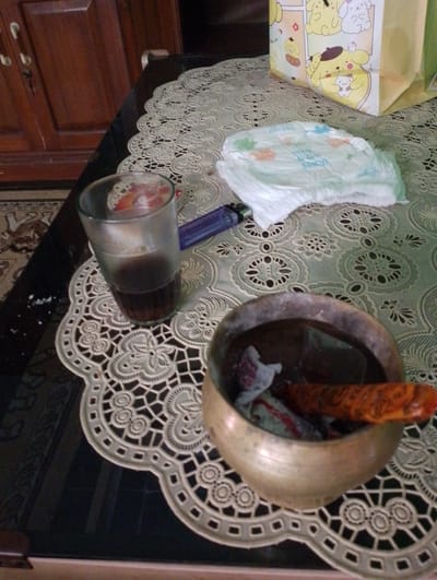 The image depicts a table with a lace doily. On the doily, there is a glass containing a dark liquid, a lighter, a diaper, and a brass bowl containing some items. The background shows a wooden cabinet and a rug. The scene appears to be indoors, likely in a residential setting in Sukamanah, Indonesia. There are no people visible in the image.