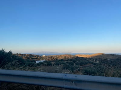 The image displays a coastal landscape in Φάρος, Greece, under a clear, gradient blue sky. In the immediate foreground, a dark grey metal guardrail runs horizontally across the bottom of the frame, partially obscuring dry, light-brown grasses and dark green shrubs. The midground features rolling hills covered extensively in dense, dark green low-lying vegetation, consistent with olive groves, intermingled with patches of terraced, lighter-brown earth. Two white, rectangular buildings are visible within this terrain: one larger structure partially hidden by foliage on the left-center, and a smaller, distinct building positioned further to its right. The background reveals the expansive, calm Aegean Sea, with several hazy, distant islands or landmasses discernible on the horizon. Sunlight, originating from a low sun angle (consistent with morning or late afternoon), illuminates the landscape, casting prominent shadows to the left across the hills. No human figures, animals, or explicit actions are present in the scene.