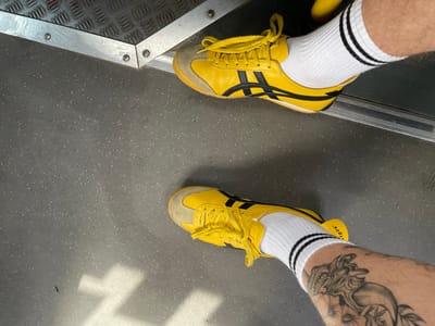 The image shows a pair of yellow Onitsuka Tiger sneakers with white socks that have black stripes. One shoe is in the foreground and the other is slightly further back and to the left. The shoes are on a gray floor with small white flecks. There is a metal step or threshold in the upper left corner of the image. The right leg of the person wearing the shoes has a tattoo of a dragon on the calf. The scene appears to be indoors, possibly on a bus or train, given the metal flooring and the general appearance of the environment. The location is not specified in the image itself, and thus Rechytsa, Belarus cannot be confirmed.