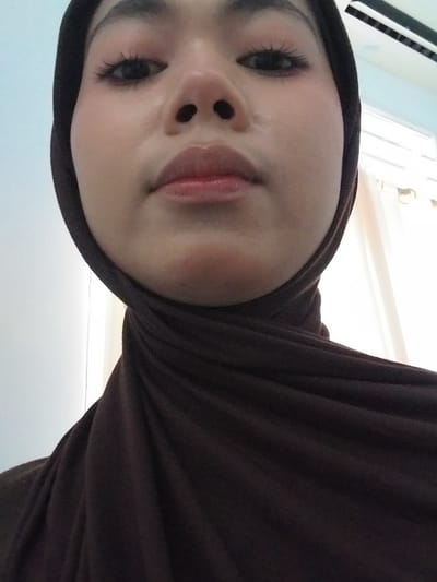 A close-up, low-angle shot shows the face of a person wearing a dark brown hijab. The subject is looking directly at the camera. Their eyes are open with visible eyelashes and subtle eye makeup. The nose is centered, and the lips are slightly parted, appearing to have a light gloss or lipstick. The skin tone is light. The background is out of focus, showing a light blue wall on the left and a white curtained window on the right, with what appears to be a ceiling fan visible at the top. The image was taken in Watampone, Indonesia.