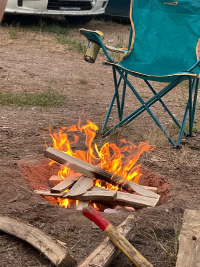 The image shows a campfire in a natural setting, likely outdoors. The fire is burning brightly with flames licking at several pieces of firewood. To the left of the fire is a teal camping chair with orange trim, its legs extending into the dry, brown earth and sparse grass. A white vehicle, possibly a van, is partially visible to the far left.
To the right of the fire, a wooden tool with a red and yellow handle, resembling a shovel or axe, rests on the ground. Additional pieces of wood are scattered around the campfire. The ground appears to be dirt with dry vegetation and some green grass interspersed.
The lighting suggests it might be daytime, possibly late afternoon or early evening, given the warmth of the fire and the light on the objects. The overall scene conveys a sense of outdoor recreation, perhaps camping or a gathering around a fire. There is no visible text in the image except for "ACTIWELL" on the camping chair. The setting appears rustic and possibly remote, consistent with a rural or wilderness location. No people are visible in the image.