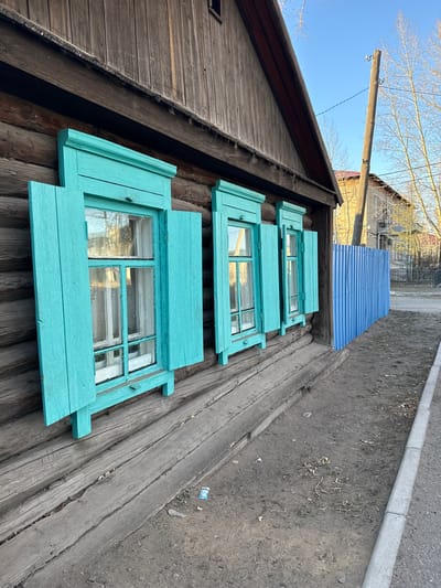 The image captures a weathered wooden house with three turquoise-framed windows in Borzya, Russia. The house is constructed of dark, aged wood logs, and its roof slopes steeply on one side. Each window has an open, turquoise shutter. Beyond the house, a blue metal corrugated roof is visible, and in the background, a street or path is adjacent to a strip of dirt and vegetation. A wooden utility pole stands tall against a clear sky with bare tree branches. The photograph is taken from a low angle, emphasizing the height and age of the building.