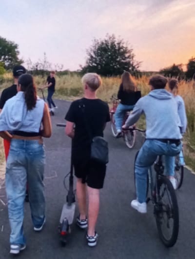 A group of seven individuals is depicted moving along a paved asphalt path in Brzezina, Poland. The ambient lighting suggests either dusk or dawn, with the sky displaying a gradient of pale orange, pink, and light blue. The path is bordered by dry, tall grasses and scattered foliage, with a prominent deciduous tree visible in the mid-background.

In the foreground, from left to right:
1.  A female, viewed from the rear, with dark hair pulled back, dressed in a light grey cropped hoodie, a dark undergarment, light blue baggy jeans, and white sneakers. She is walking.
2.  A male, viewed from the rear, with short blonde hair, wearing a black t-shirt, black shorts, and black and white sneakers. He carries a black sling bag and is pushing a grey electric scooter.
3.  A male, viewed from the rear, with dark hair, wearing a light grey hooded sweatshirt, light blue jeans, and white sneakers. He is astride a dark-colored bicycle.

Further back on the path, from right to left:
1.  A female, viewed from the rear, with long dark hair, wearing a dark top and blue jeans, riding a red bicycle.
2.  A female, partially obscured, with light hair, also on a bicycle.
3.  A male, largely obscured behind the foreground individuals, wearing a dark cap and dark t-shirt.
4.  A male, positioned further back and to the left, wearing a dark top and blue jeans, operating a second scooter.

All subjects are proceeding in the same general direction away from the camera's perspective.