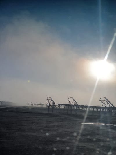 A bright sun shines over a desolate, flat landscape. Rows of dark, skeletal structures, resembling industrial equipment, stretch into the distance under a hazy sky. Raindrops or dust particles are visible in the foreground, partially obscuring the view. The overall impression is one of starkness and desolation.