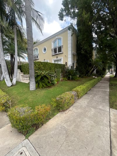 A sidewalk runs alongside a lush green hedge. A two-story building with a beige facade is visible behind the hedge. The building features arched windows, decorative balconies, and ornate trim. Tall palm trees and other foliage surround the building. A large tree provides shade overhead. The scene suggests a residential area with well-maintained landscaping.