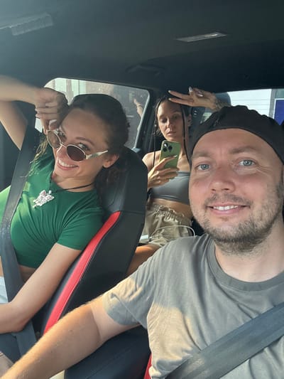 A rotated, bright selfie captures three smiling individuals inside a vehicle, likely a car. In the foreground, a man with a beard and blue eyes, wearing a black beanie and a grey t-shirt, smiles directly at the camera. He is secured by a seatbelt.
Behind him, a woman with braided dark hair, wearing a grey crop top and light-colored shorts, holds a green smartphone. Tattoos are visible on her left arm, and she is looking upwards, possibly at the man or the camera.
In the background, a third woman with curly dark hair and round, tinted sunglasses smiles broadly. She is wearing a green t-shirt and a prominent butterfly-shaped necklace, also secured by a seatbelt.
The scene is illuminated by bright sunlight, casting shadows on the subjects and the car's interior, indicating daytime. The interior features black and red upholstery on the car seats and a dark headliner. All three individuals appear cheerful and relaxed, suggesting a casual, happy moment during travel. No text is visible within the image. The provided location context is Hà Giang 1 Ward, Vietnam.