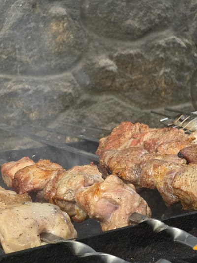 This is a close-up, vertically oriented, eye-level shot of meat grilling on skewers. The subjects are pieces of meat, likely pork, threaded onto metal skewers. The meat is browning and appears to be seasoned with pepper. Smoke is visible rising from the meat, indicating it is actively cooking.
The background on the left is a textured, grey stone surface, possibly the wall of a fireplace or an outdoor grill. The right side of the image shows the dark, textured surface of a grill, with the spiral shape of the skewers clearly visible. The overall scene is one of food preparation, likely a barbecue or cookout.
There are no people visible in the image. The lighting suggests it could be daytime, but the exact time of day is not discernible. The weather is also not indicated. There is no visible text. The location context is Pidhorodne, Ukraine, which suggests this could be a traditional Ukrainian dish being prepared outdoors. The focus is entirely on the food and the grilling process.