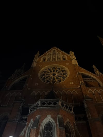 The image shows the exterior of the Co-cathedral of St. Peter and Paul in Osijek, Croatia, at night. The co-cathedral, constructed of red brick, is lit up, highlighting its Gothic Revival architectural details. The most visible feature is the large rose window located on the front facade. The sky is black.