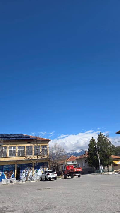 The image captures a street view in Slatino, Bulgaria, featuring a clear blue sky on the left. On the right side of the frame, there is a street with parked vehicles: a white SUV and a red tractor. A two-story building with solar panels on its side is situated along the street. The building's facade displays advertisements. In the background, there are houses, trees, and mountains partially obscured by clouds.