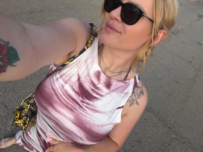 A woman with blonde hair pulled back, wearing large dark sunglasses, a tie-dye dress, and a designer handbag, stands on a paved surface in Ayia Napa, Cyprus. She is holding her right arm up, taking a selfie. She has tattoos on both arms. The background is a gray, pebbled surface, likely a road or sidewalk. The lighting suggests it is daytime, possibly late afternoon due to the warm tones.