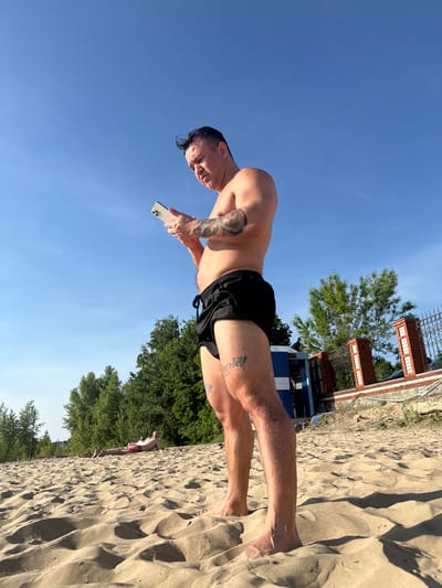 A man stands on the sandy beach in Matiushino, Russia, holding a cell phone. He is wearing black swim trunks. The sky is clear and blue. Trees and a building structure are in the background.