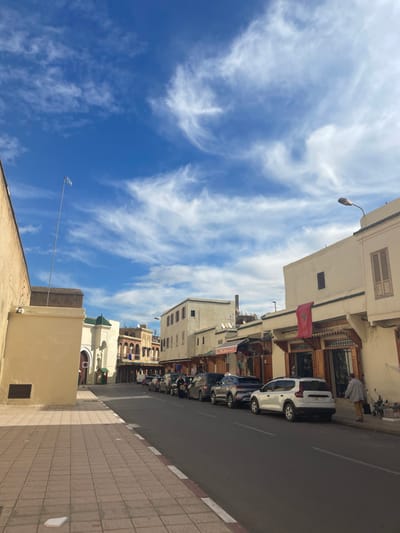 The image displays a street scene within Mechouar Fes Jdid, Morocco, under a bright blue sky with scattered white cirrus clouds. A wide, tiled sidewalk, featuring light and dark brown square tiles, occupies the left foreground, bordered by a plain, light-colored wall. Behind this wall, a tall, slender metallic pole extends vertically. Further down the street on the left, a building with a white facade and a distinctive green-tiled domed roof is visible.
The dark asphalt road, marked with a white line on its left edge, recedes into the distance, populated by a continuous line of parked vehicles along its right side. These include multiple SUVs and sedans in various colors, such as black, grey, and white, with a white Dacia Duster prominently positioned in the mid-ground.
On the right side of the street, a series of multi-story buildings with cream-colored facades line the thoroughfare. Many ground-floor units function as shops, characterized by ornate wooden storefronts and awnings. A red Moroccan flag, featuring a green star, hangs horizontally above one such shop entrance in the mid-ground. A street light with a single lamp arm is mounted on a building in the upper right quadrant. Multiple satellite dishes are visible on the rooftops of these structures.
One individual, clad in a light-colored, long garment, walks away from the viewer on the right-side sidewalk, moving towards the background. Other indistinct figures are visible further down the street among the parked cars and shops. Shadows cast on the left sidewalk and under vehicles indicate direct sunlight from the right side of the frame.