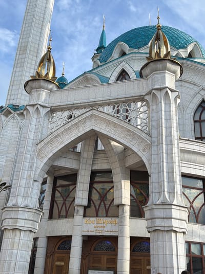 The image shows a close-up, exterior view of a white stone building with intricate architectural details, possibly a mosque given the presence of a minaret and a domed roof structure. The building features elaborate stonework, including decorative carvings, arches, and columns. A tall, slender minaret with a golden finial rises prominently on the left side of the frame. A turquoise-colored dome is visible in the upper left quadrant. The building's facade is adorned with geometric patterns and Arabic script carved into the stone. The windows appear to have stained glass elements.
The lighting suggests it is daytime, with sunlight casting shadows on the facade, indicating a clear or partly cloudy sky. The overall impression is one of grandeur and ornate craftsmanship.
Visible text on the building includes golden lettering in what appears to be Cyrillic and possibly Arabic script. One sign reads "Кол Шәриф", which translates to "Kul Sharif" in Tatar and Russian, a strong indicator that this is the Kul Sharif Mosque in Kazan, Russia, a famous landmark. The text suggests it is a public building, likely a religious or historical site. There are no people clearly visible in this frame. The scene is static, focusing solely on the architectural elements of the building against a sky background.