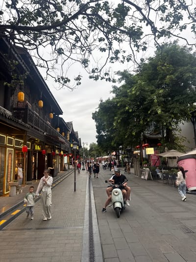 This image captures a bustling pedestrian street in Kuanzhai Alley, Qingyang District, Chengdu, China, under an overcast sky. The street is lined with traditional two-story Chinese buildings featuring dark wood facades, intricate carvings on balconies, and tiled roofs. Numerous yellow lanterns hang from the eaves of the buildings on the left, with some red lanterns also visible at ground level.

On the left side of the street, storefronts display traditional wooden signs and some modern neon lighting, including one prominent sign with Chinese characters and another appearing to be "伍杏". A person in traditional attire stands inside one of the shop entrances near the foreground. In the immediate foreground, a woman wearing a white shirt and striped pants holds the hand of a young boy in a light-colored jacket as they walk towards the viewer. The woman is drinking from a cup.

The street itself is paved with stone blocks in a herringbone pattern, interrupted by sections of square tiles and a decorative metal drainage grate running down the center. Further down the street, a man wearing a helmet and a black t-shirt is riding a light-colored scooter with another person on the back.

On the right side of the street, large, leafy green trees provide shade, some with their branches extending across the upper portion of the frame. Below the trees, more shops and eateries are visible, some offering outdoor seating under beige umbrellas. A sign for "Molly Tea" (茉莉茶) is discernable among the storefronts. Numerous pedestrians populate the street, walking in both directions, contributing to the lively atmosphere. The sky is a uniform light grey, indicating either a cloudy day or early evening.