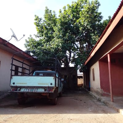 In Mangu, Nigeria, a pale blue pickup truck is parked on a dirt road between two buildings. The truck has a metal cage over its bed. The building on the left has a light-colored wall and a red corrugated metal roof. A satellite dish is mounted on the roof. The building on the right has a dark red facade and a red corrugated metal roof. A large tree with green leaves stands behind the truck. The sky is clear and blue.
